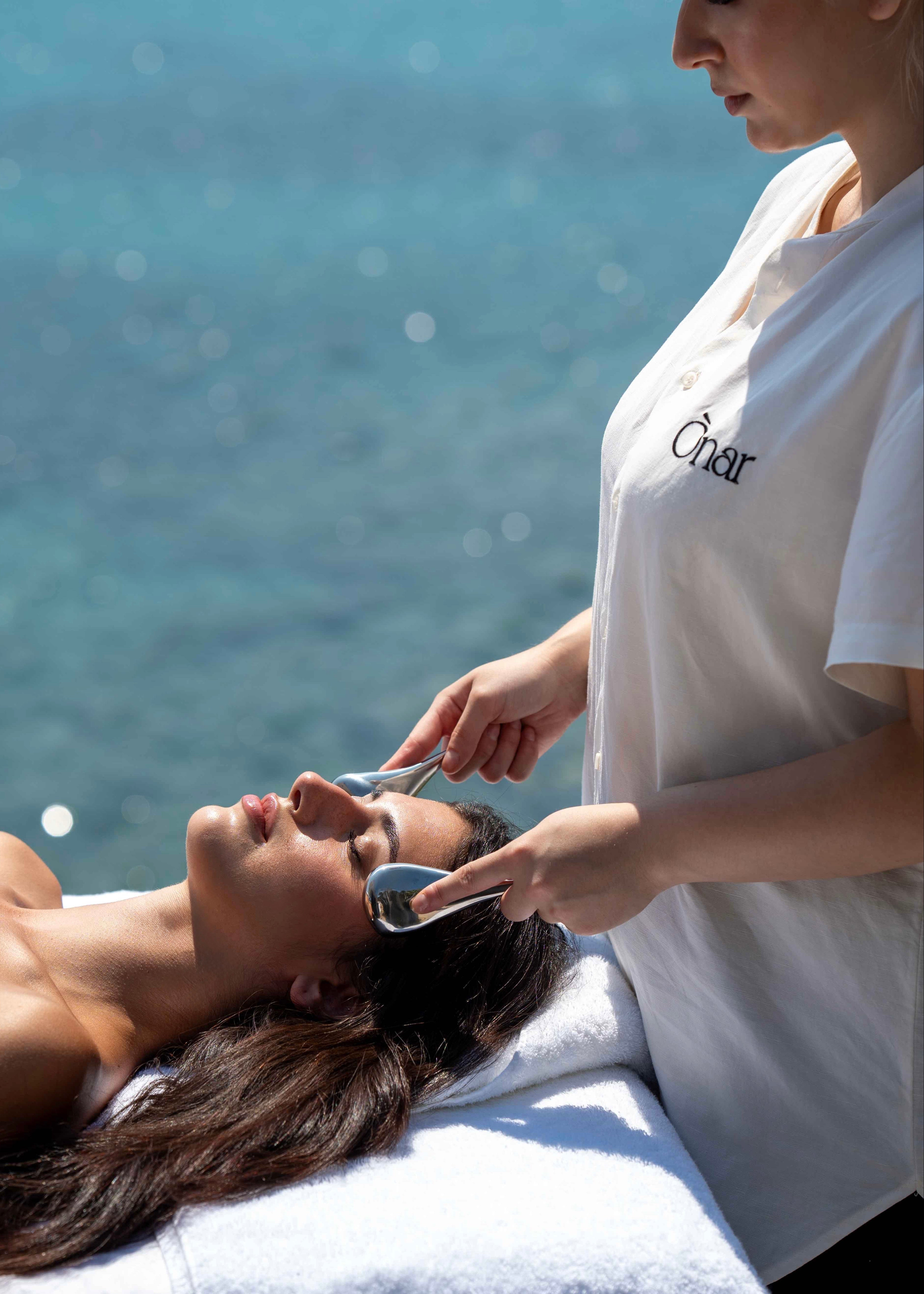 A spa treatment scene by the sea with a therapist applying a facial mask to a relaxed client lying on a white treatment table. The therapist wears a white ONAR shirt, and the background shows a bright Mykonian blue sea.